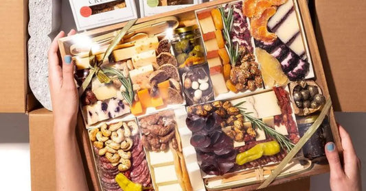 Gourmet charcuterie gift box with cheeses, nuts, fruits, and meats tied with a gold ribbon inside packaging