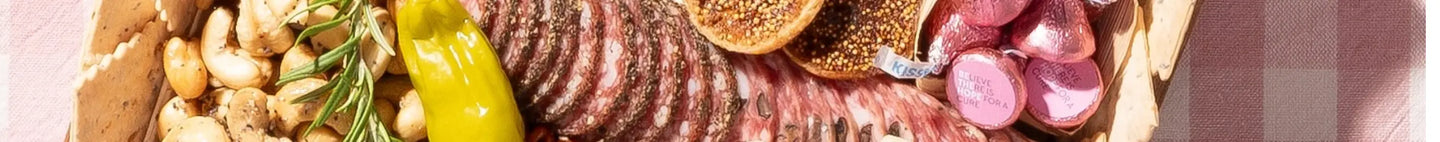 Charcuterie board with cured meats, cashews, figs, pepperoncini, rosemary, and pink foil chocolates.