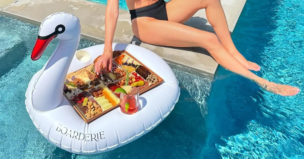 Charcuterie board floating on a swan pool float beside a woman dipping her feet in the water.