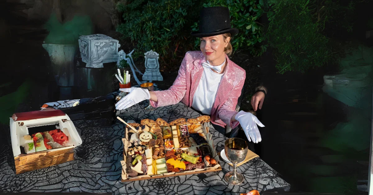 Halloween snack board with themed cheeses, meats, and treats served on a spooky table by a costumed hostess.
