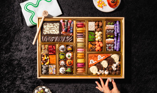 Boarderie's Halloween Dessert Board featuring festive Halloween macarons, chocolate-dipped pretzels, candy corn, ghost truffles, and spooky decorated treats arranged in a gold gift box