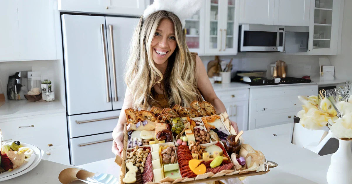 Charcuterie board for parties held by a smiling woman in a bright kitchen, filled with meats, cheeses, and festive touches.