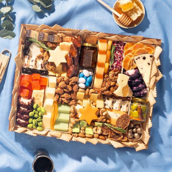 Hanukkah cheese board with Star of David cheeses, nuts, dried fruit, and honey on blue linen.
