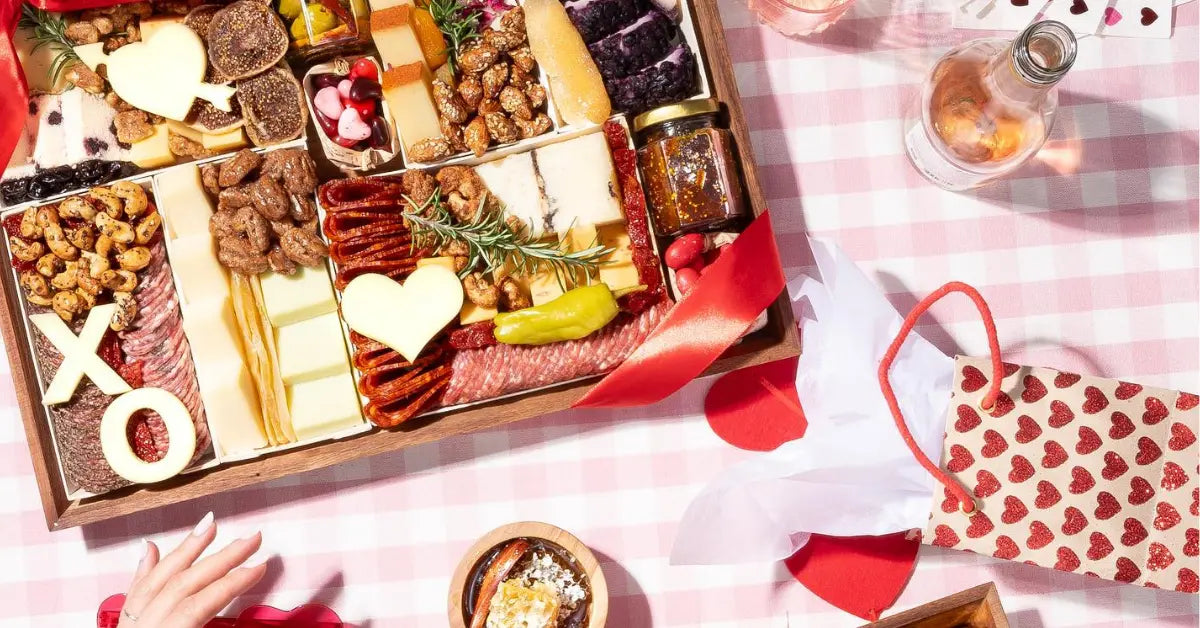 Valentine's Day charcuterie board with heart-shaped cheese, meats, nuts, and sweets styled on a pink gingham picnic setup.