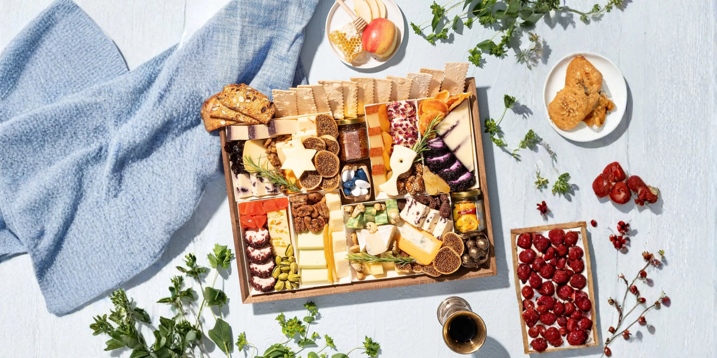 Charcuterie board delivery in a wooden gift tray filled with artisan cheeses, dried figs, candied nuts, dried fruit, crackers, and gourmet accompaniments, styled on a light blue surface with fresh herbs, raspberries, and honeycomb.