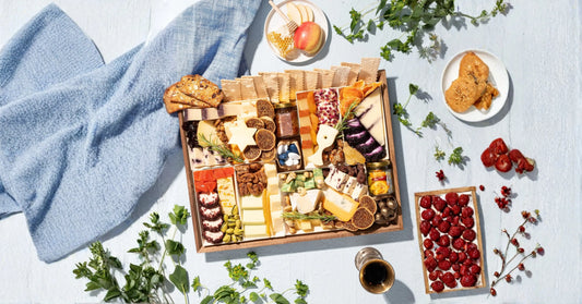 Charcuterie board delivery in a wooden gift tray filled with artisan cheeses, dried figs, candied nuts, dried fruit, crackers, and gourmet accompaniments, styled on a light blue surface with fresh herbs, raspberries, and honeycomb.