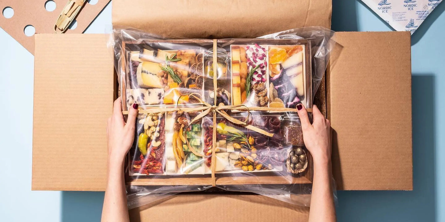 charcuterie board delivery unboxed by two hands lifting a ribbon-tied board packed with meats, cheeses, and nuts from a shipping box