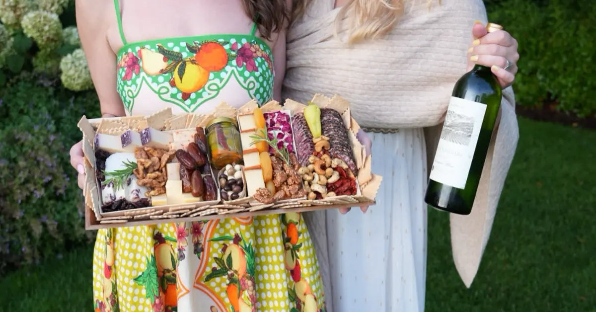 Charcuterie board for beginners held outdoors by two women, featuring simple cheeses, cured meats, nuts, and wine.