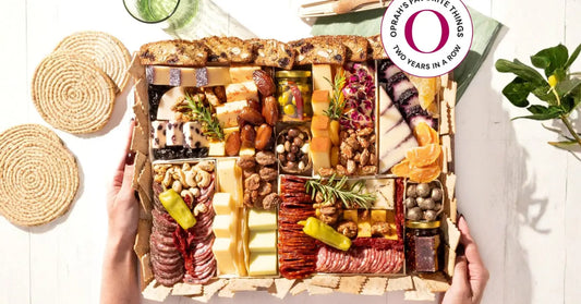 Overhead artisanal charcuterie board with assorted cheeses, cured meats, nuts, dried fruit, crackers, and garnishes on a bright table.
