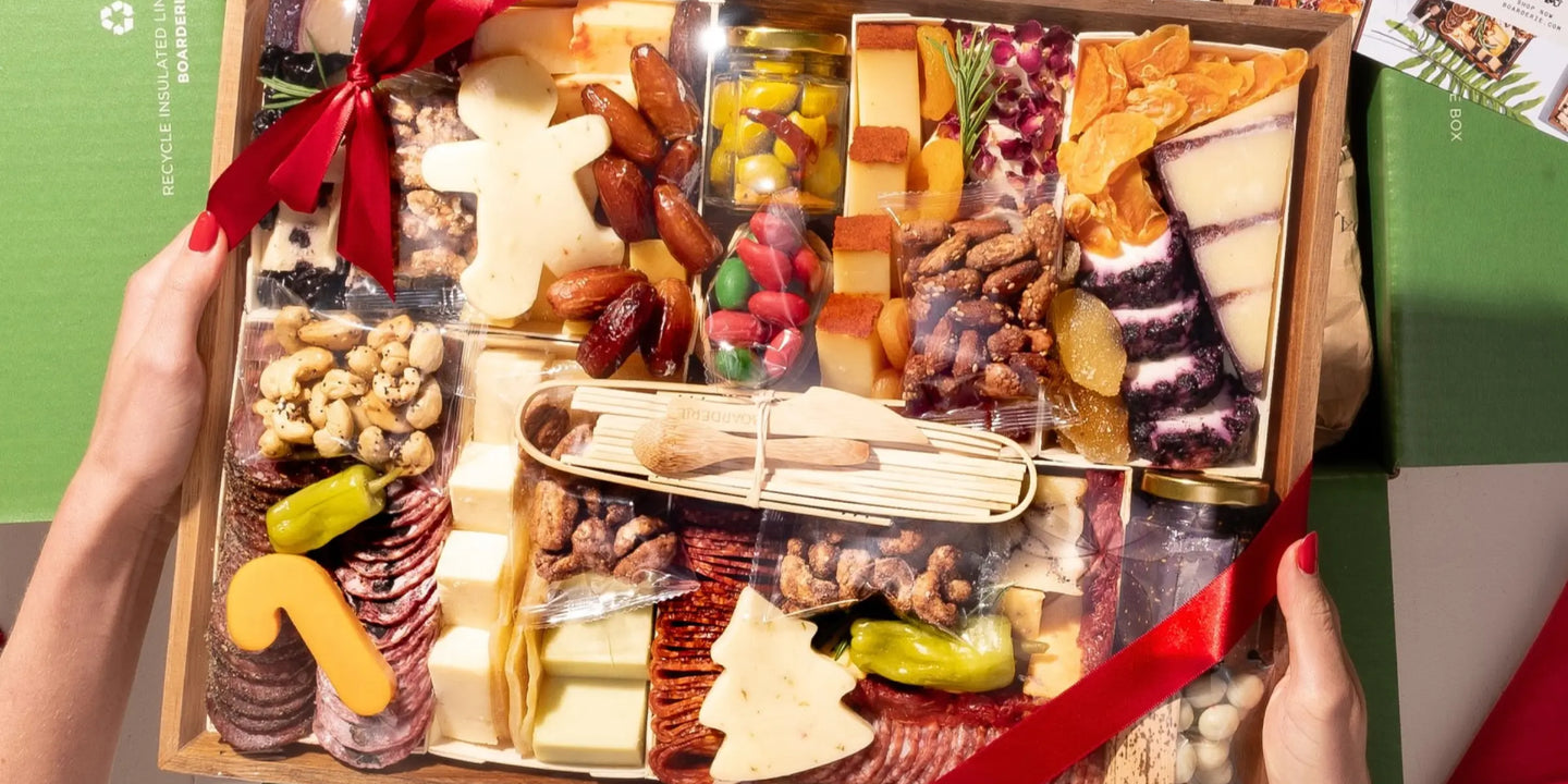 classic charcuterie board with meats, tree-shaped cheese, nuts, and dried fruits wrapped in a red ribbon inside a holiday green box