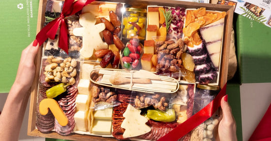 classic charcuterie board with meats, tree-shaped cheese, nuts, and dried fruits wrapped in a red ribbon inside a holiday green box