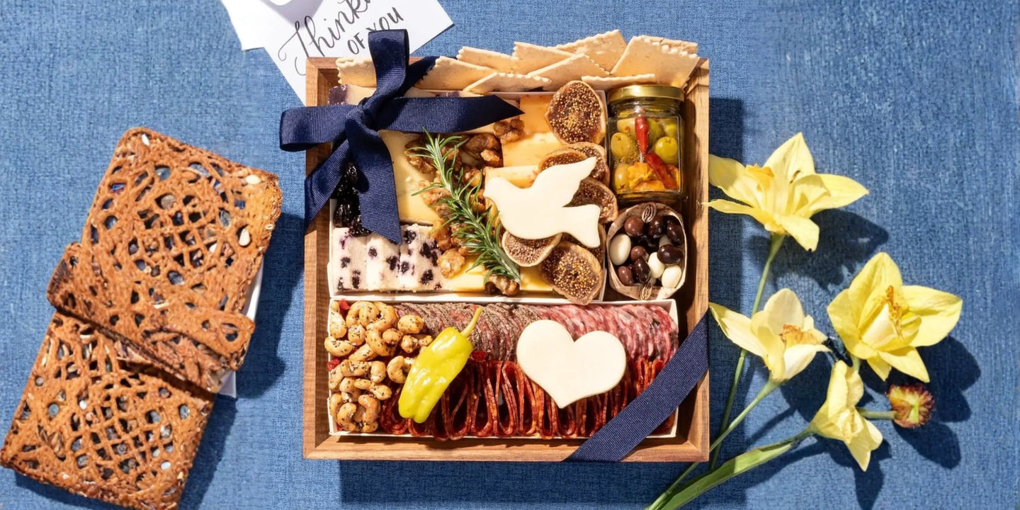 condolence food baskets: Condolence charcuterie box with cheeses, sliced salami, crackers, nuts, olives, and heart-shaped cheese accents tied with a navy bow