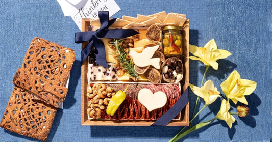 condolence food baskets: Condolence charcuterie box with cheeses, sliced salami, crackers, nuts, olives, and heart-shaped cheese accents tied with a navy bow