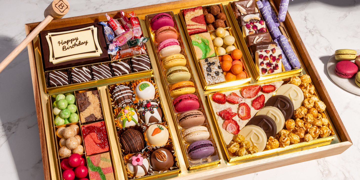 Birthday dessert board with macarons, chocolates, popcorn, and a 'Happy Birthday' plaque, set on a marble table.