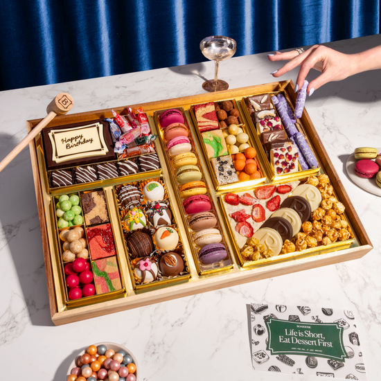 Birthday dessert board with macarons, chocolates, popcorn, and a 'Happy Birthday' plaque, set on a marble table.