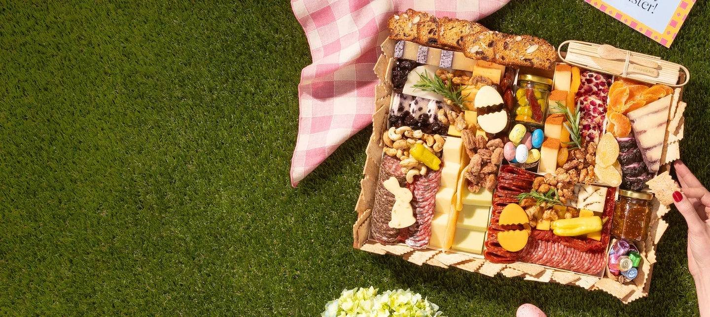 Summer charcuterie board arranged on grass with cheeses, colorful candies, nuts, and meats beside a picnic blanket.