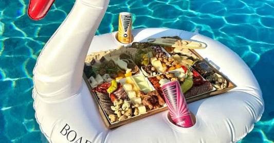Charcuterie board with cheeses, meats, and nuts floating in a pool on a white swan float with drinks.