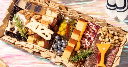 Festive cheese board with “2025” cutout, dates, olives, nuts, and artisan cheeses arranged with crackers and garnishes