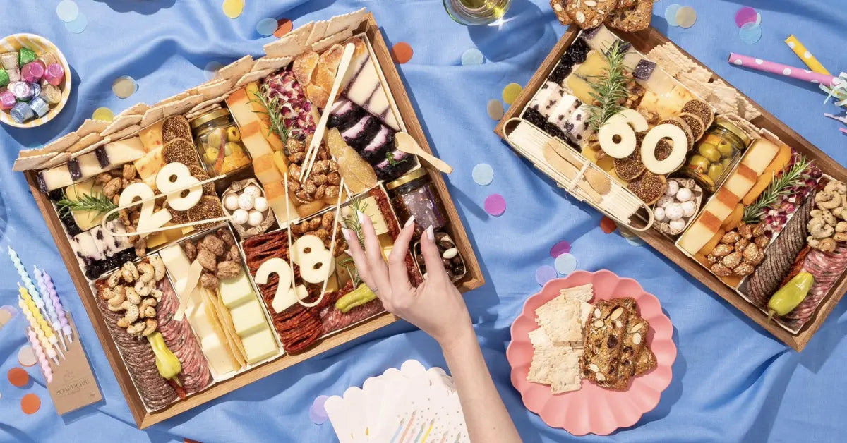 Birthday charcuterie boards from Boarderie customized with ages in cheese, displayed on a blue tablecloth with confetti and candles.