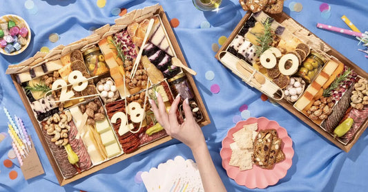 Sweet and savory charcuterie board with birthday numbers, cheese, meats, nuts, and chocolate on a festive blue backdrop.