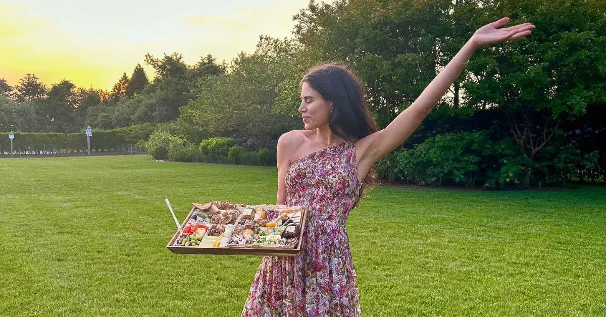 Woman in a floral dress holding a gourmet cheese tray with meats, cheeses, and fruits in a lush outdoor setting.