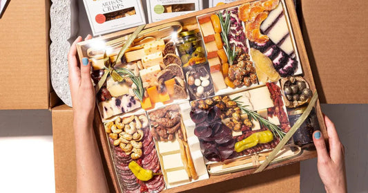 Meat and cheese platter wrapped in plastic with assorted cheeses, salami, nuts, and dried fruits inside a shipping box.