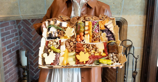 Modern charcuterie board with autumn-themed cheese cutouts, meats, nuts, and fruits held in front of a cozy fireplace.