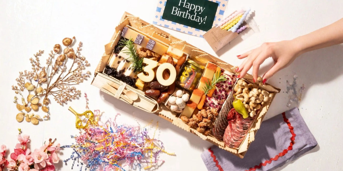 Birthday-themed appetizer board with “30” cheese numbers, crackers, salami, nuts, and a hand reaching in beside a Happy Birthday card and candles