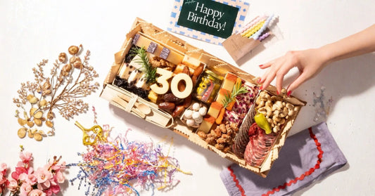Birthday-themed appetizer board with “30” cheese numbers, crackers, salami, nuts, and a hand reaching in beside a Happy Birthday card and candles