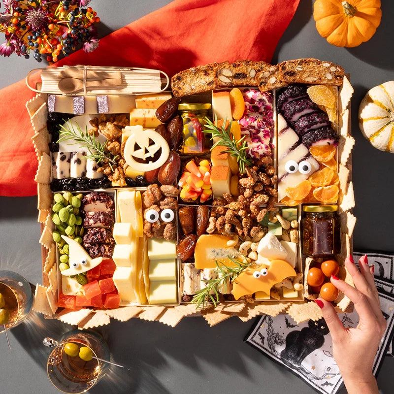 Halloween-themed charcuterie board with spooky decorations, a jack-o'-lantern cheese cutout, googly eyes on cheeses, and candy corn on an orange backdrop.