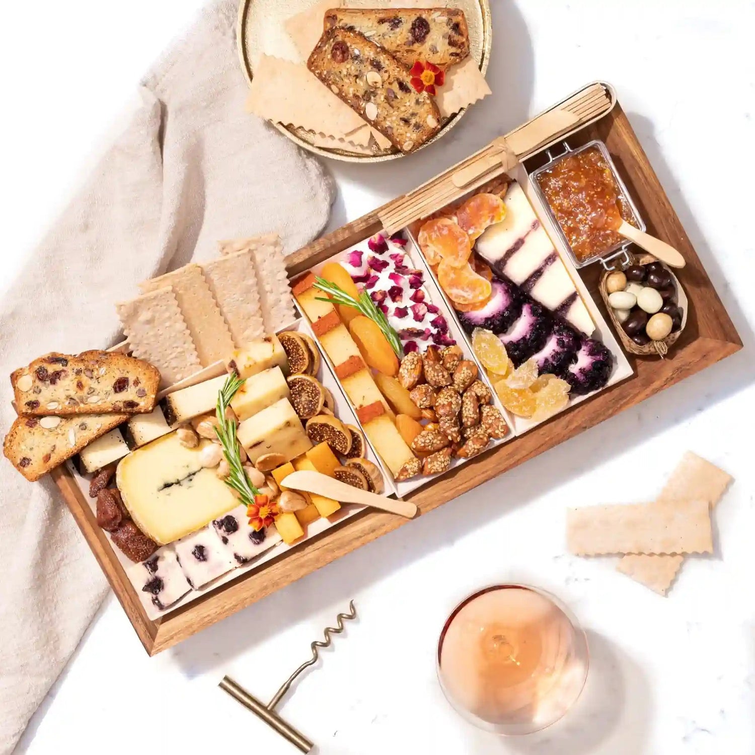 Wooden charcuterie board with assorted cheeses, dried fruits, crackers, jam, and nuts, accompanied by a glass of wine and bread slices.