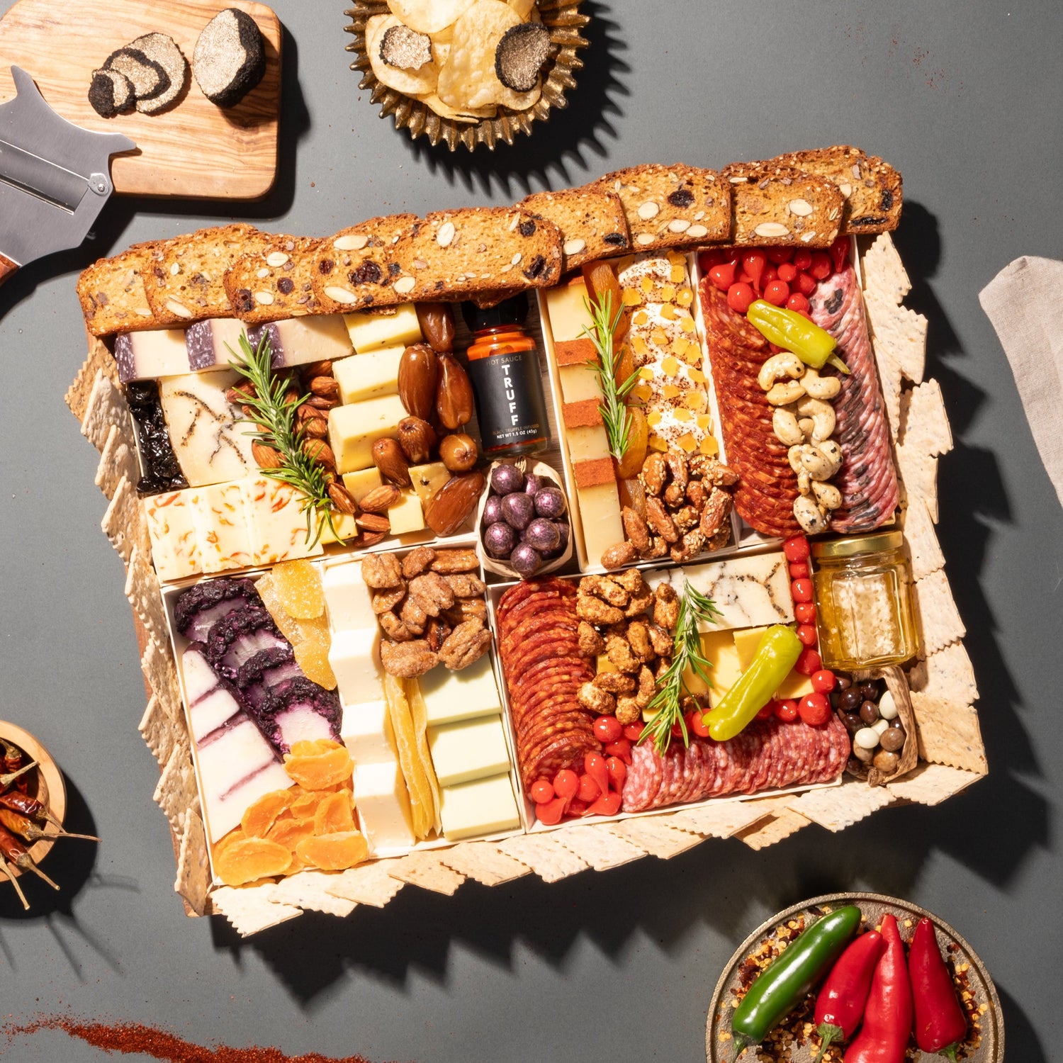 Charcuterie board with an array of cheeses, meats, nuts, truffle hot sauce, and crackers, garnished with peppers on a dark surface.