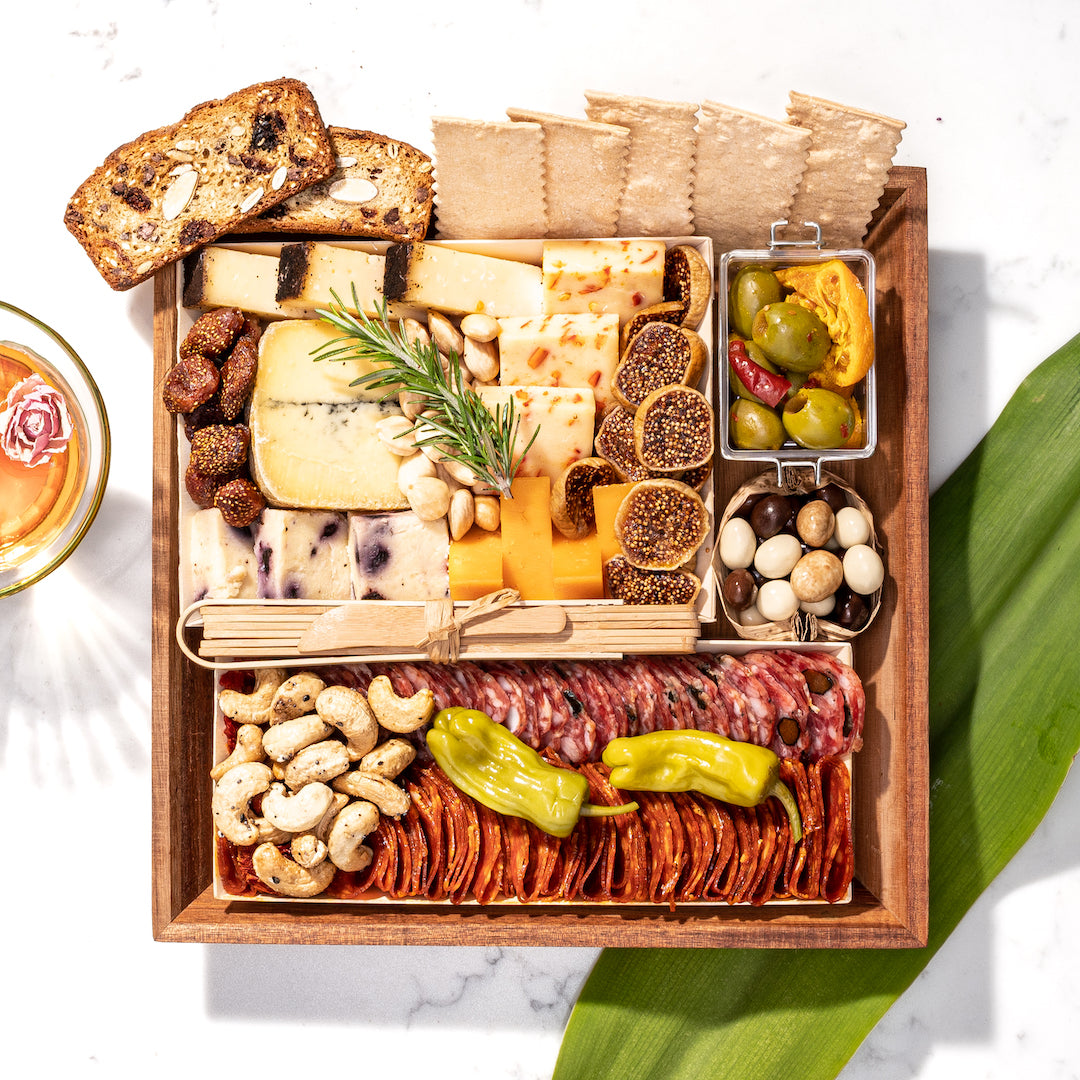 Wooden charcuterie board with assorted cheeses, meats, nuts, olives, figs, crackers, and bread slices on a white marble surface.