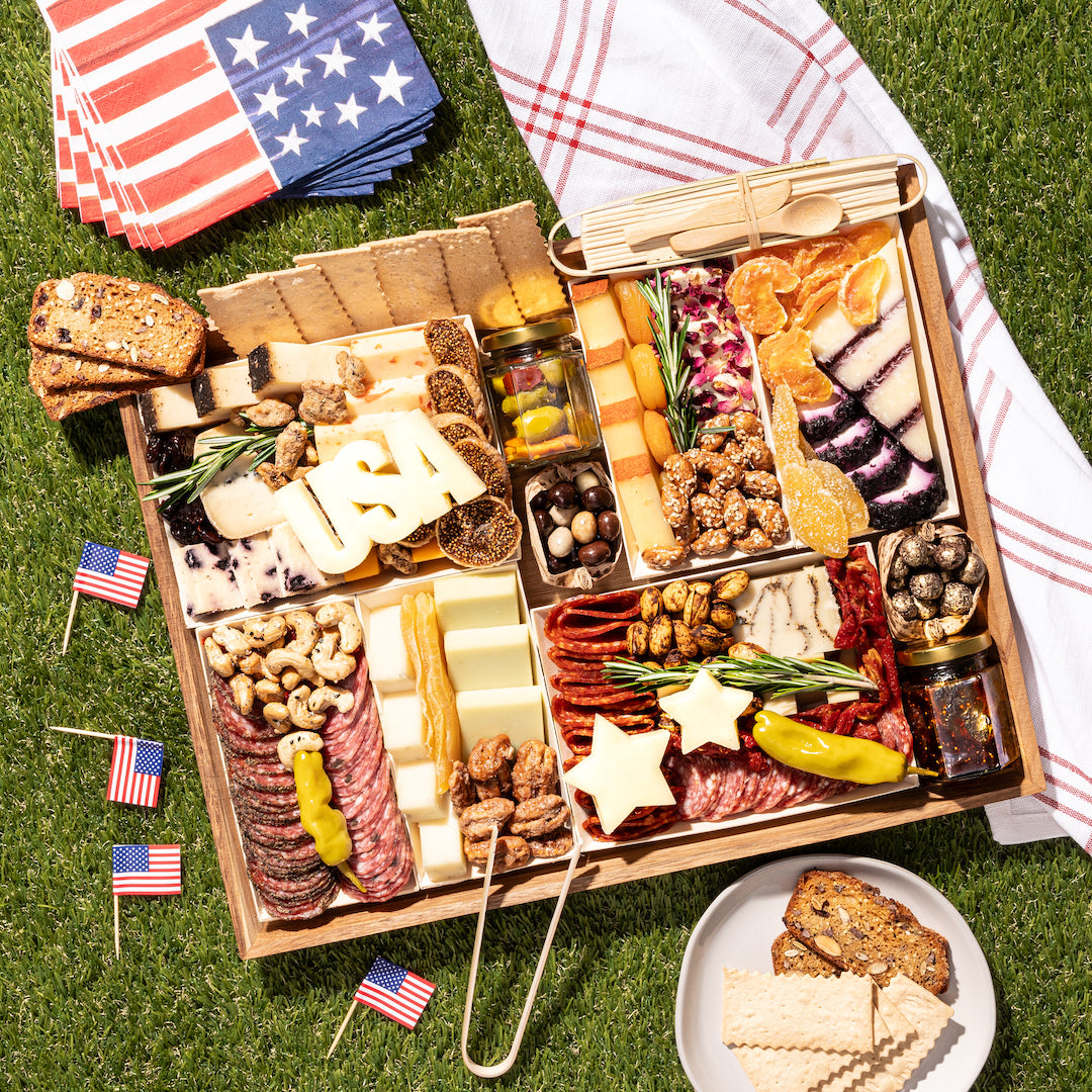 Patriotic charcuterie board with "USA" and star-shaped cheeses, meats, and snacks, decorated with American flags on grass.