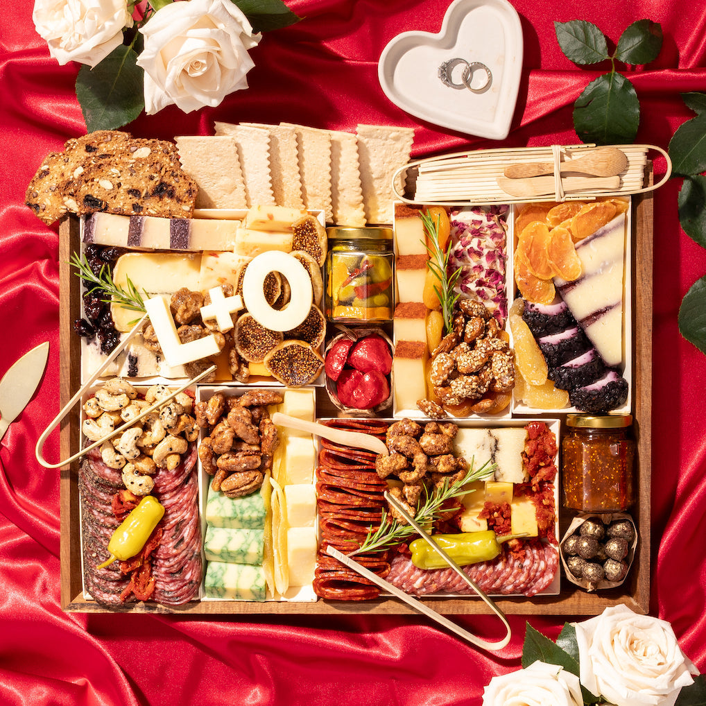 Romantic charcuterie board with "L+O" cheese initials, meats, cheeses, and nuts, set on red satin with roses and wedding rings.