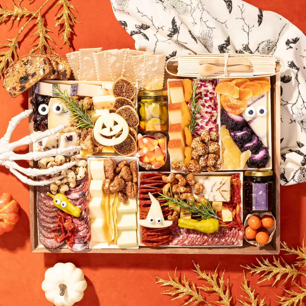 Halloween-themed charcuterie board with spooky decorations, a jack-o'-lantern cheese cutout, googly eyes on cheeses, and candy corn on an orange backdrop.