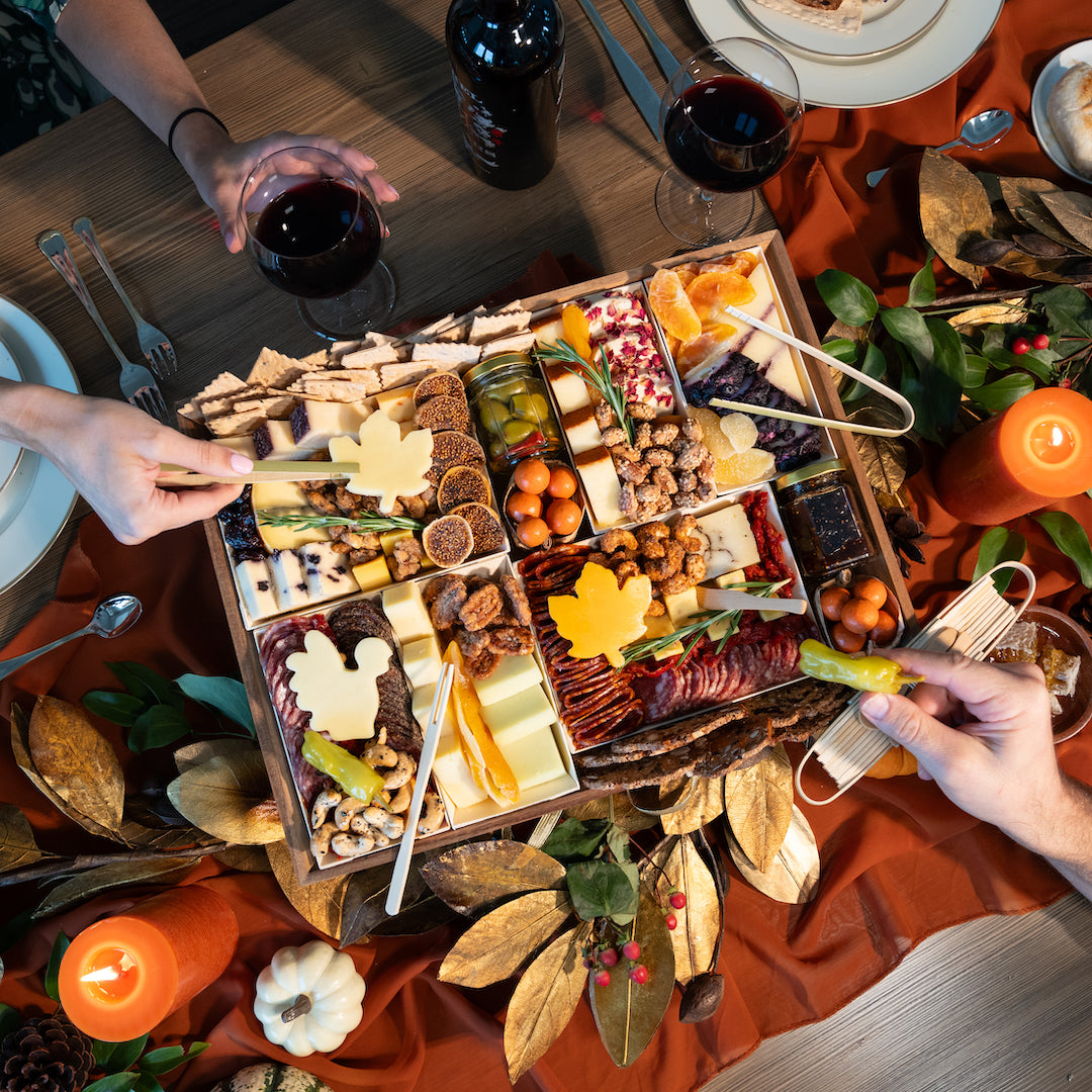 Thanks Giving-themed charcuterie board with leaf-shaped cheeses, meats, fruits, and nuts, surrounded by candles and fall decor on a festive table.