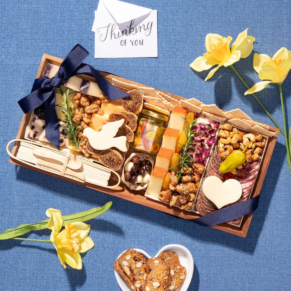 Sympathy charcuterie board with cheeses, nuts, figs, and a heart-shaped cheese piece, set with yellow flowers and a "Thinking of You" card.