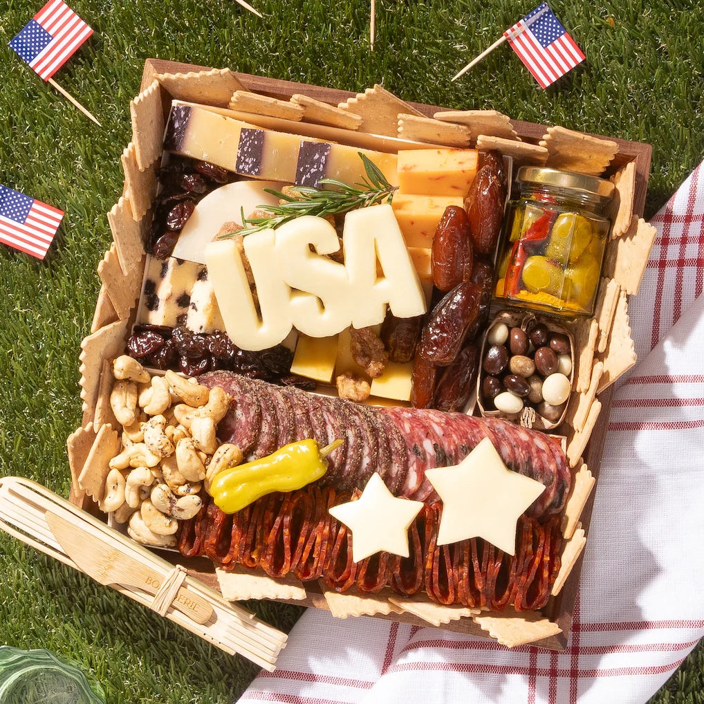 Patriotic 4th of July charcuterie board with USA-shaped cheese, star cutouts, salami, and American flags on grass.
