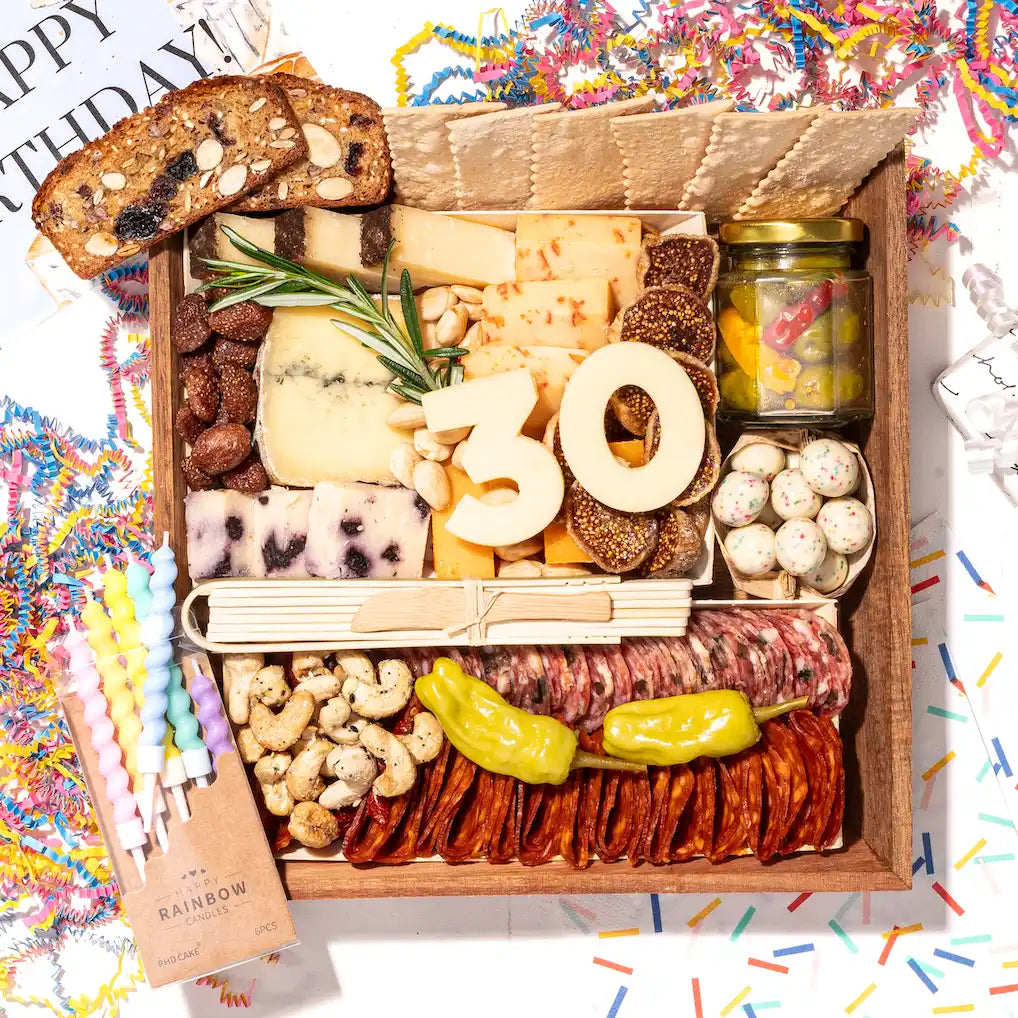 Birthday-themed charcuterie board with cheese numbers "30," meats, nuts, and crackers, surrounded by confetti and festive candles.