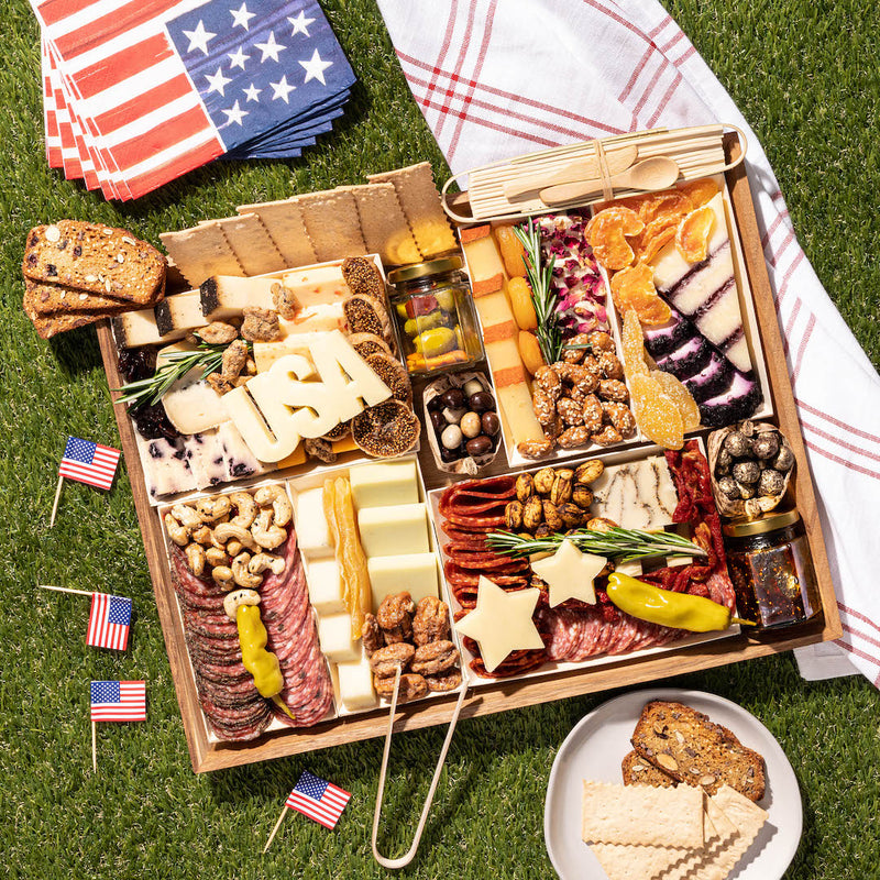 4th of July charcuterie board with patriotic-themed cheese, meats, nuts, & crackers, decorated with small American flags