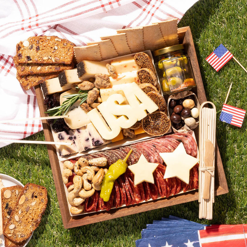 Independence Day charcuterie board with "USA" cheese letters, star-shaped slices, meats, nuts, figs, & crackers on grass