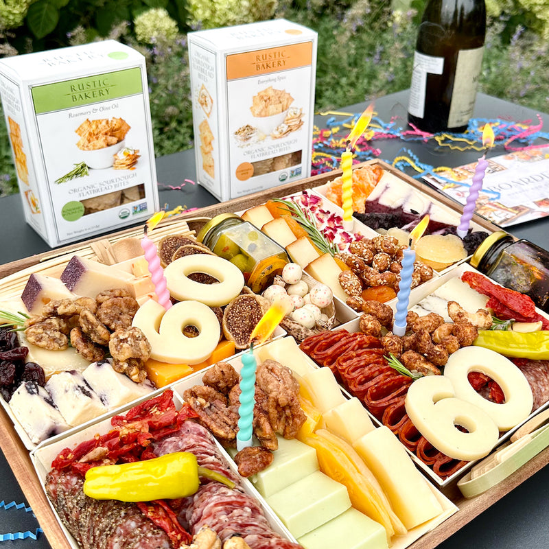 Birthday charcuterie board with "60" in cheese, colorful candles, meats, cheeses, and crackers on an outdoor table.