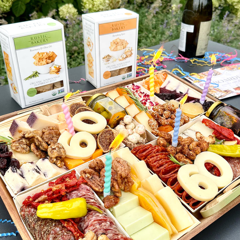 Birthday charcuterie board with "60" cheese numbers, assorted meats, cheeses, nuts, and colorful lit candles for celebration.