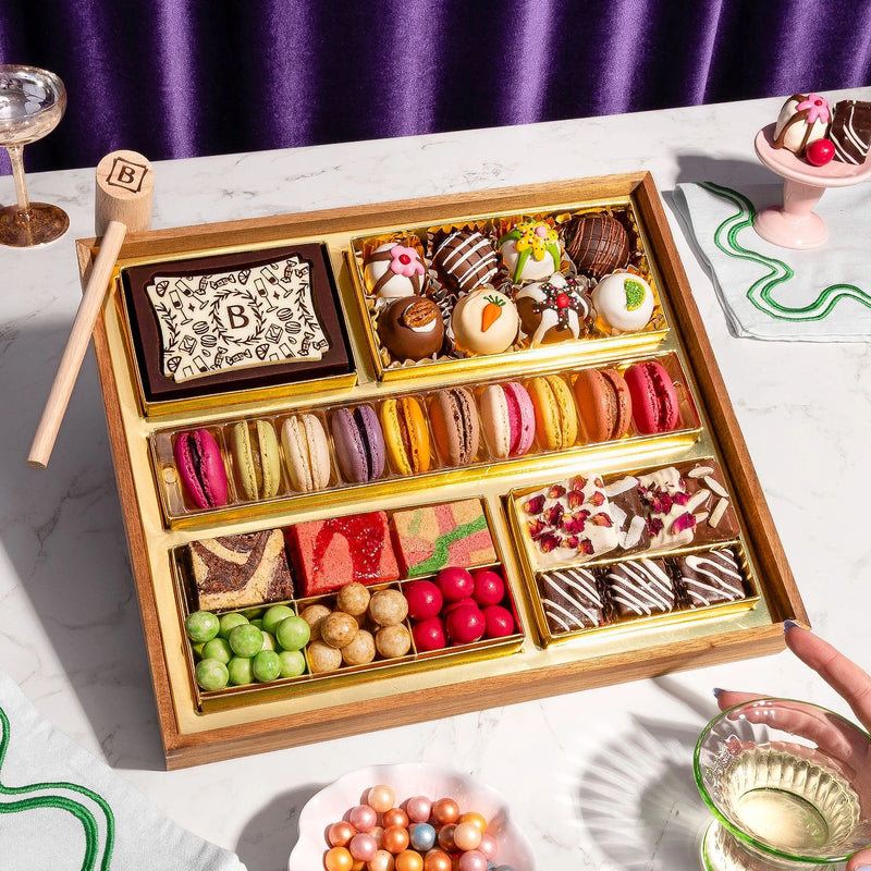 Medium chocolate dessert board featuring colorful macarons, decorated truffles, chocolate shards, gourmet candies, and fruit gummies arranged in gold compartments with a wooden smashing mallet.