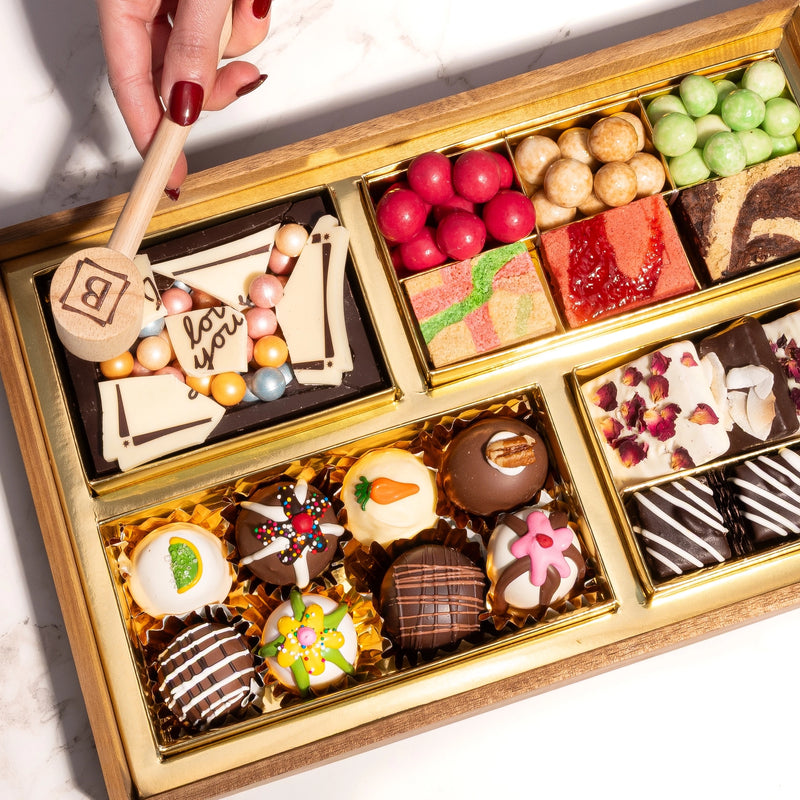 Chocolate dessert board being smashed with wooden mallet, filled with decorated truffles, macarons, candies, and fruit gummies in gold compartments.