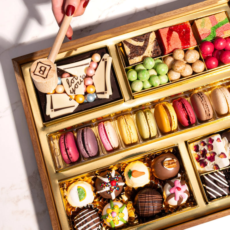 Medium chocolate dessert board being smashed with wooden mallet, displaying macarons, decorated truffles, candies, and fruit gummies in gold compartments.