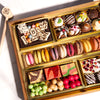 Medium chocolate dessert board featuring macarons, decorated truffles, chocolate pieces, colorful candies, and fruit gummies in gold compartments with a wooden smashing mallet.