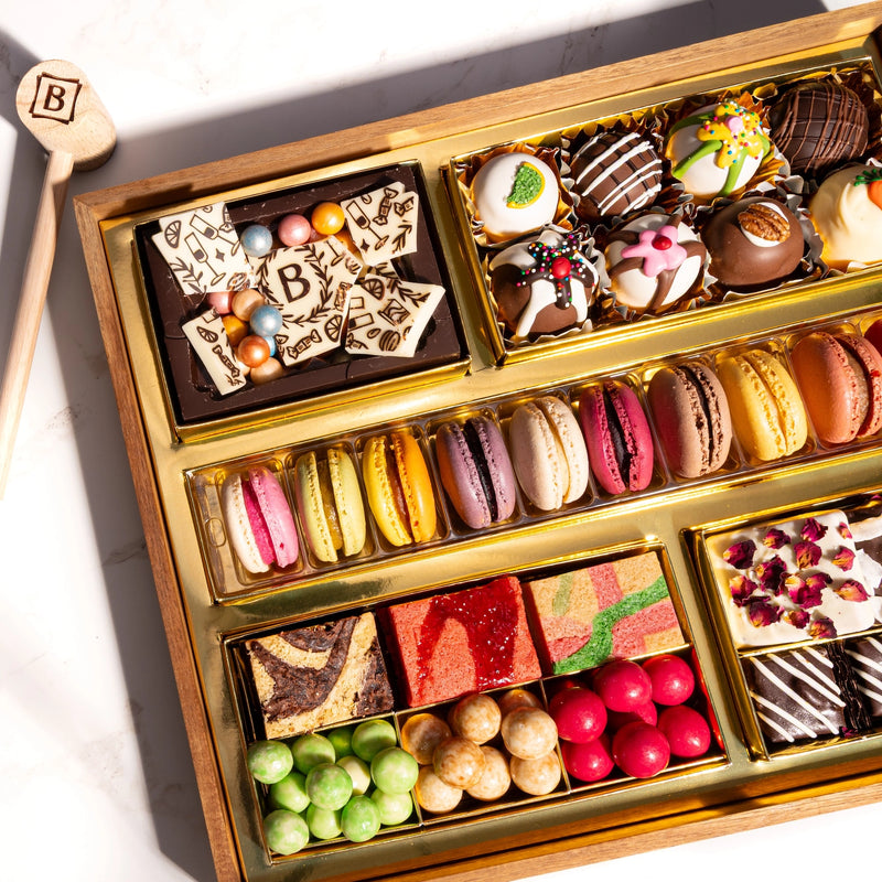 Medium chocolate dessert board featuring macarons, decorated truffles, chocolate pieces, colorful candies, and fruit gummies in gold compartments with a wooden smashing mallet.