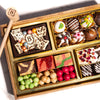 Wooden chocolate dessert board with white and dark chocolate pieces, decorated truffles, colorful candies, and fruit gummies arranged in gold compartments with a wooden smashing mallet.